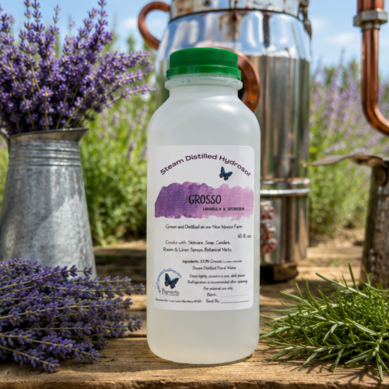A bottle of Grosso Vanilla & Stimmas steam distilled hydrosol centered on a rustic wooden table, surrounded by lavender, rosemary, a galvanized bucket, copper distillation equipment, and a blue sky background.