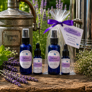 Four blue glass bottles of GROSSO handcrafted floral water and essential oil set arranged on a rustic wooden surface with lavender sprigs, a metal jug, and a clear gift bag tied with a purple ribbon. The background shows a blurred garden scene with green foliage and purple flowers.