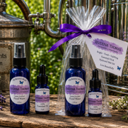 Four cobalt blue bottles of Riverina Thomas hydrosol products and a gift bag of lavender and rosemary hydrosol mist, arranged on a rustic wooden surface with metal apothecary equipment and green foliage in the background.