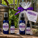 Four cobalt blue bottles of Riverina Thomas hydrosol products and a gift bag of lavender and rosemary hydrosol mist, arranged on a rustic wooden surface with metal apothecary equipment and green foliage in the background.