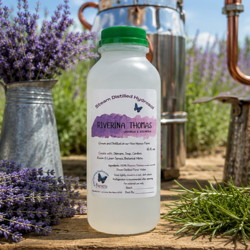 A bottle of Riverina Thomas Steam Distilled Hydrogen centered on a rustic wooden surface, surrounded by lavender sprigs, a metal pitcher, copper distillation equipment, and green rosemary in the background.