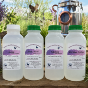 Four bottles of steam distilled herbal tinctures lined up on a wooden surface with a copper distillation pot and green foliage in the background. The bottles have white plastic bodies, white caps, and green caps on three of the bottles, and feature purple watercolor-style labels with the names Grosso, Royal Velvet, Tunstead, and Riverina Thomas.
