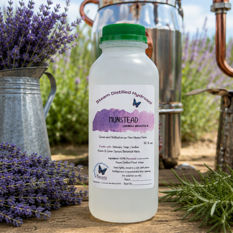 A bottle of Munstead Steam Distilled Hydracer with a green cap and white label, set on a rustic wooden surface with lavender, a metal pitcher, and copper distillation equipment in the background.