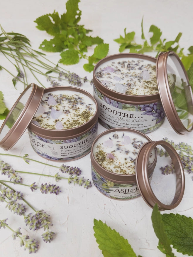 Handmade soy wax candle with lavender and mint herbs, packaged in a tin with a clear lid. The candle is surrounded by fresh mint leaves and lavender buds.