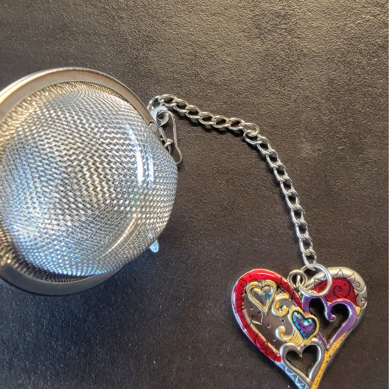 A silver-colored tea ball infuser with a decorative heart-shaped chain link attached to it, displayed against a black background.